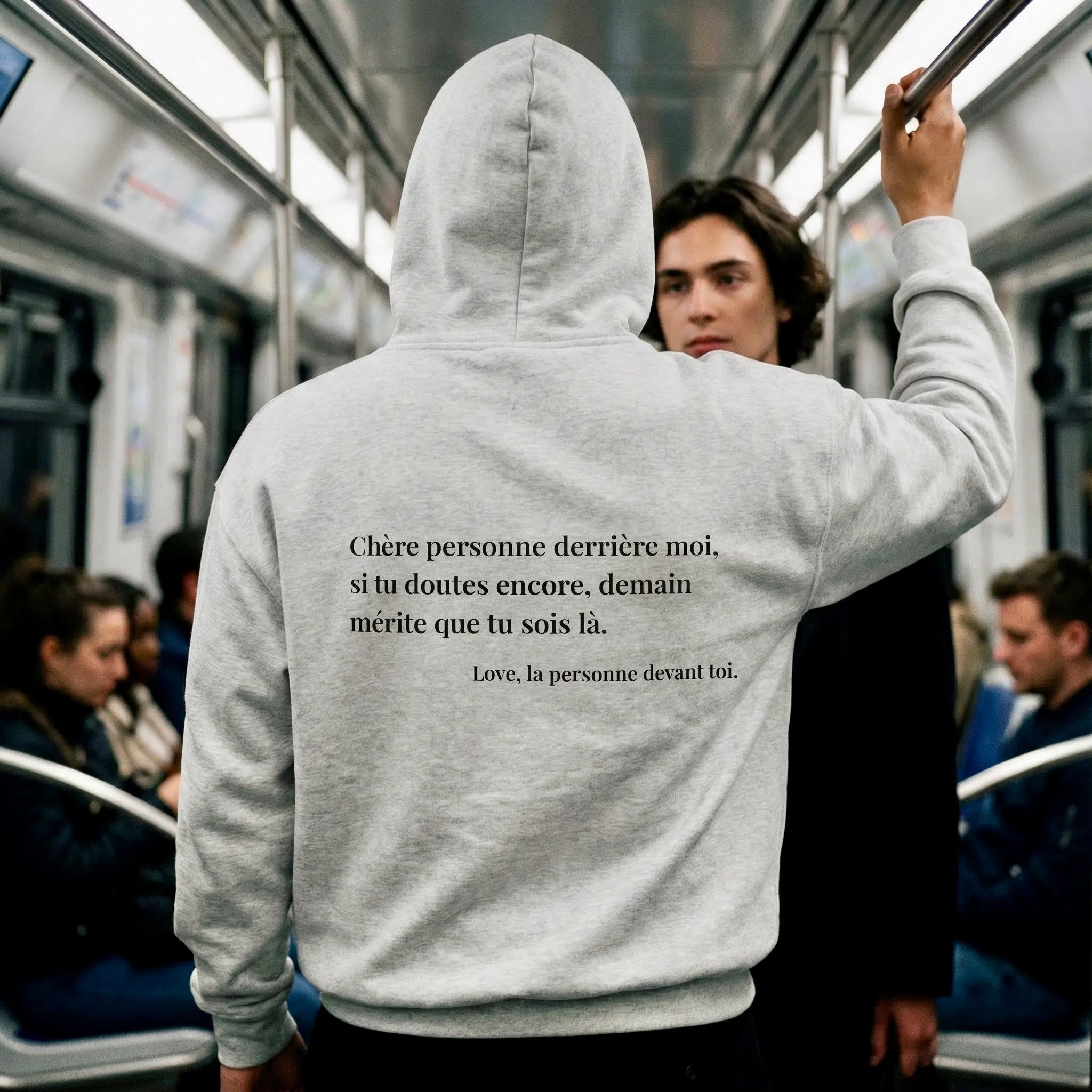 Homme de dos dans le métro portant un sweat STAY avec un message d’espoir imprimé dans le dos, lu par un passager derrière lui